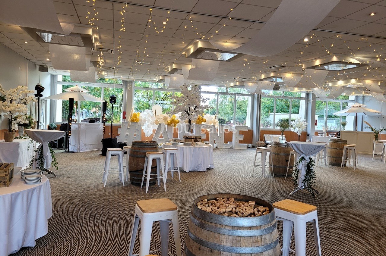 Salle de réception décorée avec des guirlandes lumineuses, tables hautes, tonneaux et grandes baies vitrées donnant sur un…