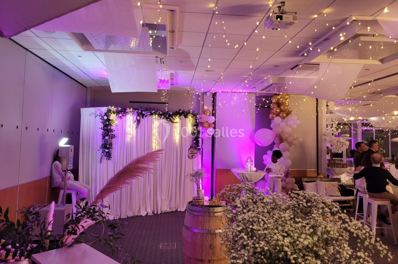 Salle décorée pour un événement avec guirlandes lumineuses, rideaux blancs, fleurs et tonneaux en bois.