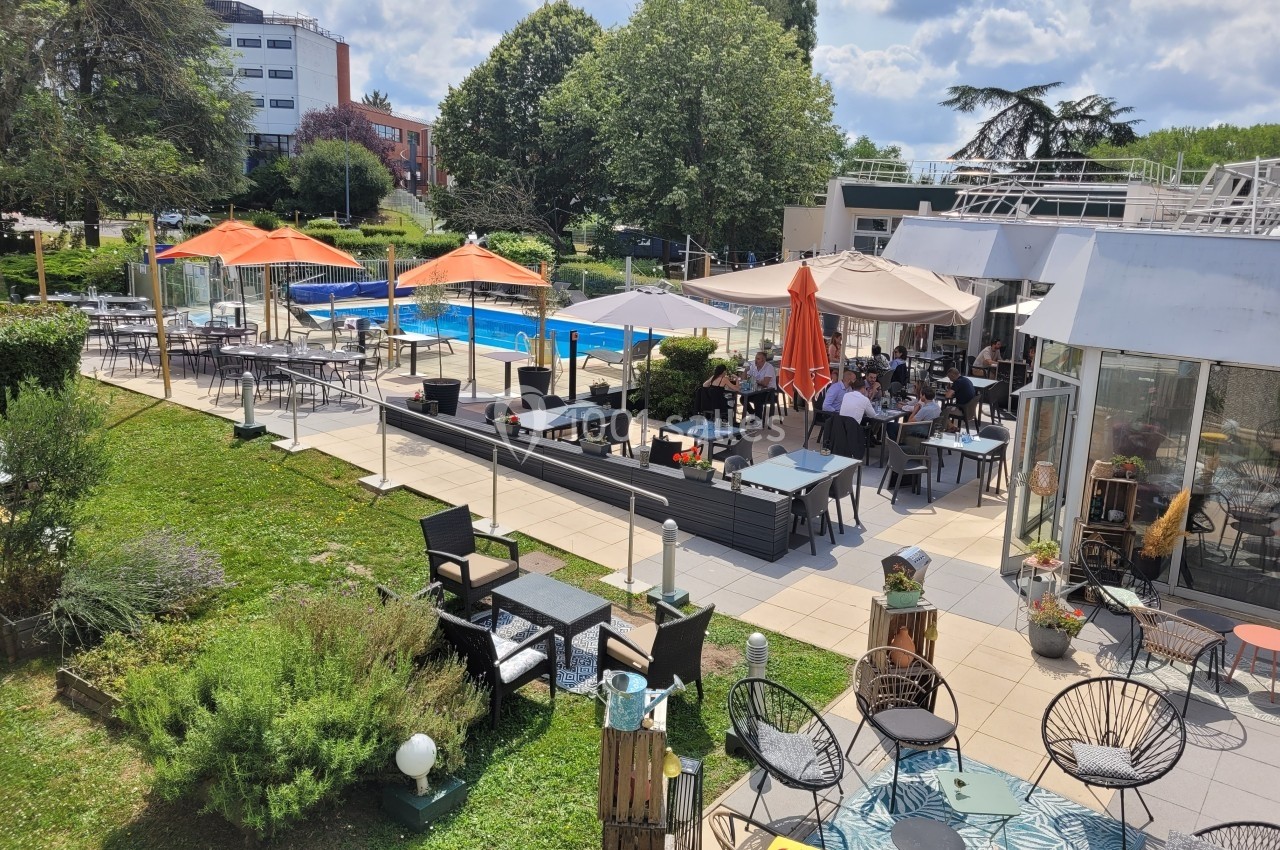 Terrasse d'un restaurant avec des tables, des parasols colorés et une piscine en arrière-plan, entourée de verdure.