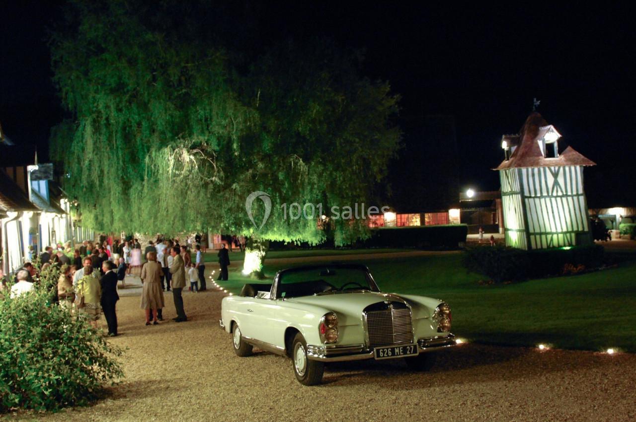 Voiture ancienne blanche garée devant un bâtiment éclairé, avec des invités rassemblés dans un jardin de nuit.