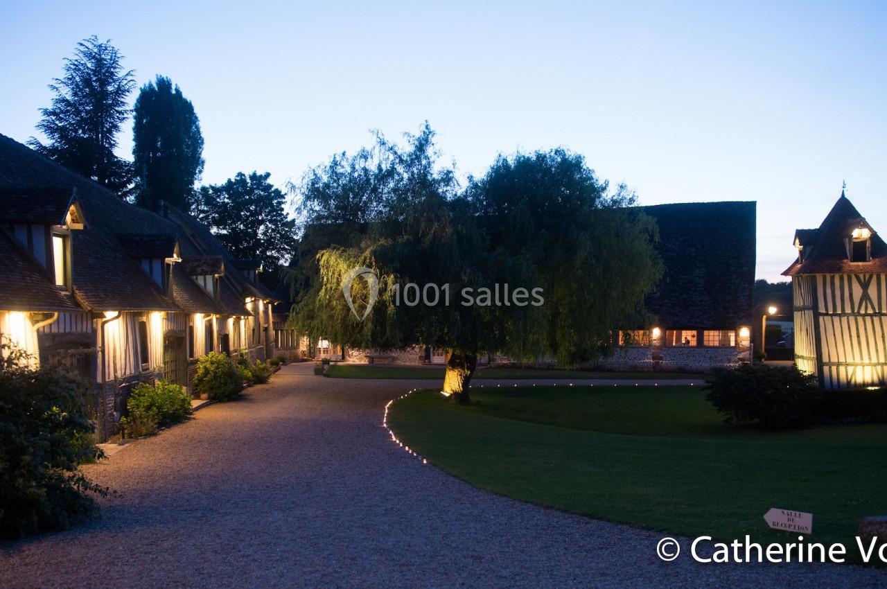 Allée éclairée bordée de bâtiments à colombages et d'un arbre au crépuscule, dans un cadre rural.