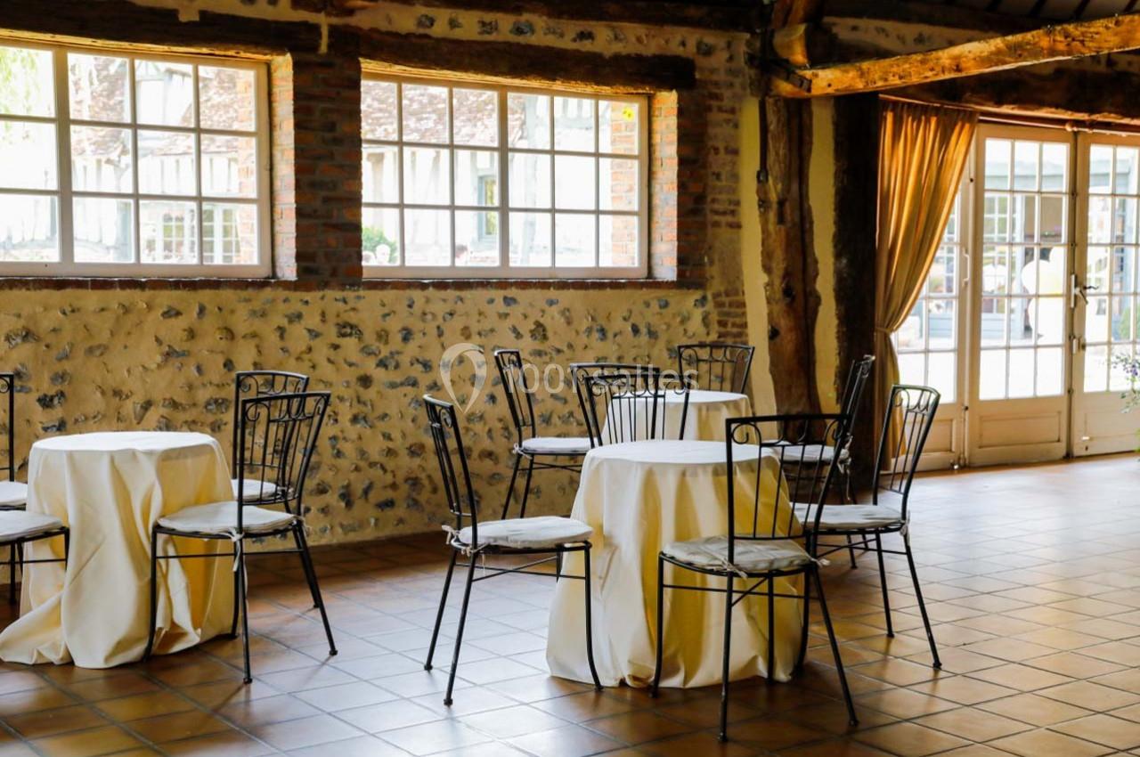 Salle lumineuse avec tables rondes drapées de nappes blanches, chaises en métal et murs en pierre apparente.