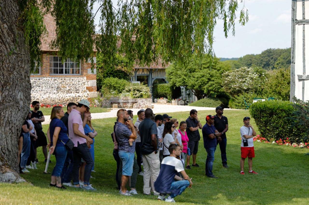 Un groupe de personnes debout sur une pelouse, écoutant une présentation en extérieur près de bâtiments et d'arbres.