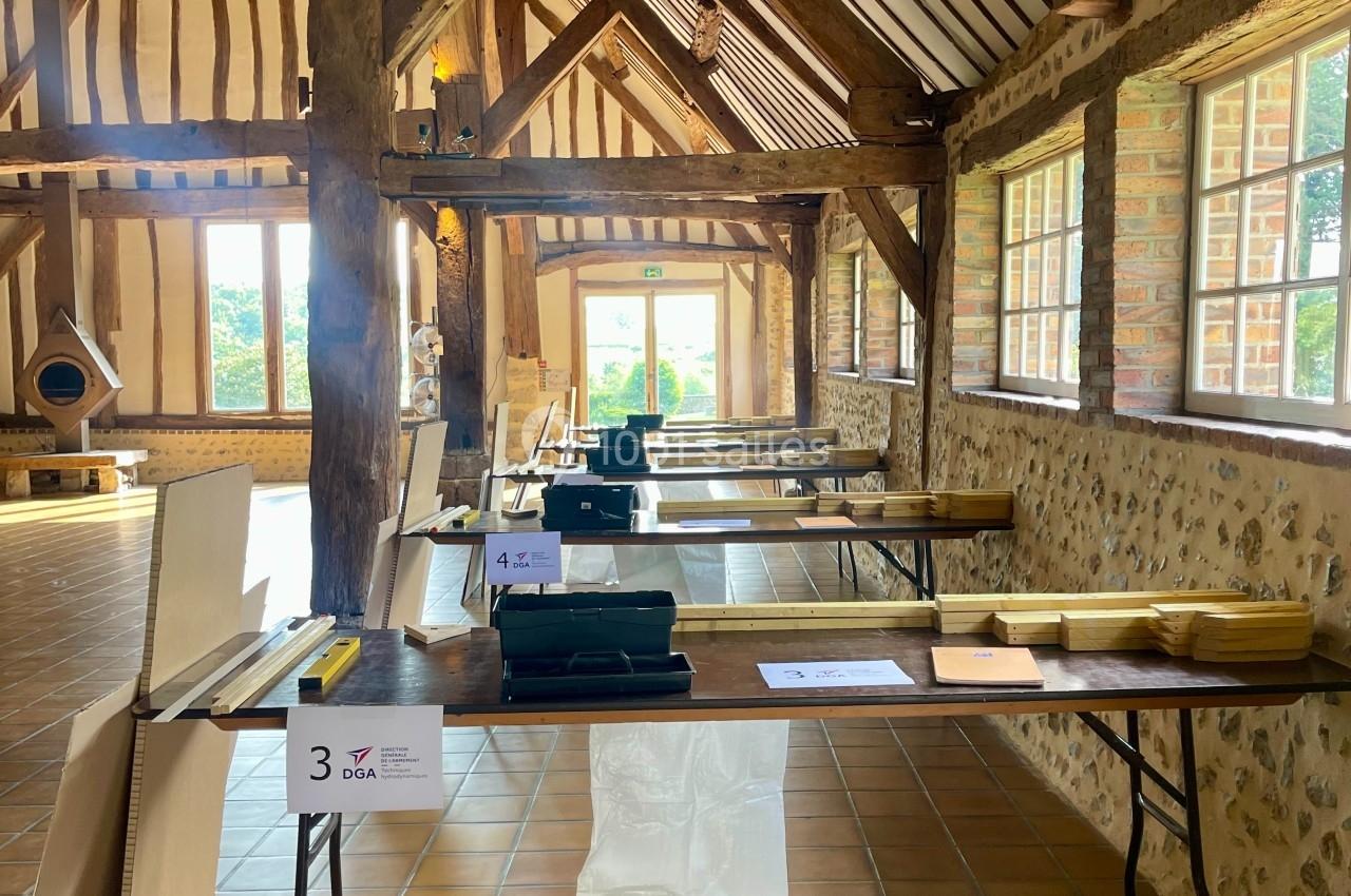 Salle en bois avec des tables alignées, des documents et des équipements, éclairée par des fenêtres et une porte ouverte.