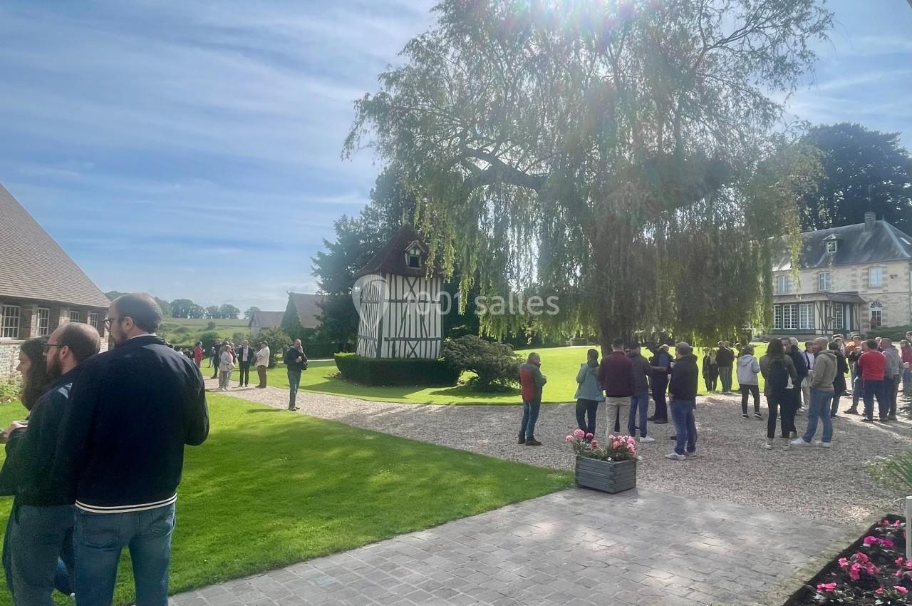 Groupe de personnes rassemblées dans une cour verdoyante avec des bâtiments traditionnels et un grand arbre sous le soleil.