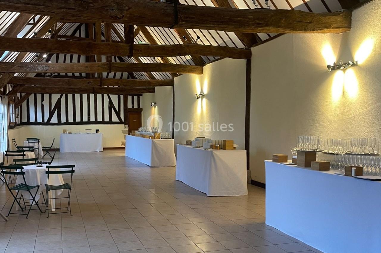 Salle avec poutres apparentes, tables dressées avec vaisselle et chaises pliantes, prête pour un événement.
