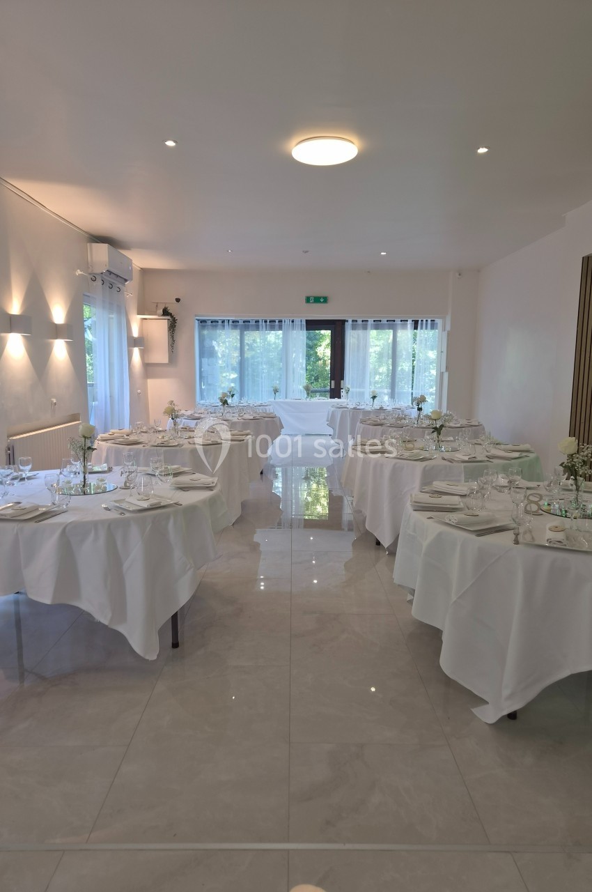 Salle lumineuse avec tables rondes dressées de nappes blanches, prête pour un événement ou un repas formel.