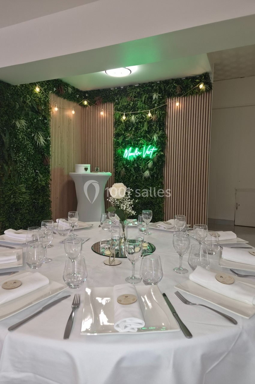 Table dressée avec vaisselle élégante, nappe blanche et centre de table floral, devant un mur végétal éclairé.