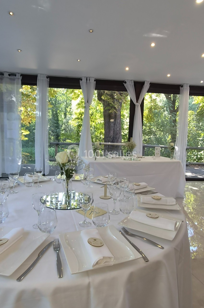 Salle lumineuse avec grandes baies vitrées donnant sur un jardin, tables dressées pour un événement avec vaisselle élégante.