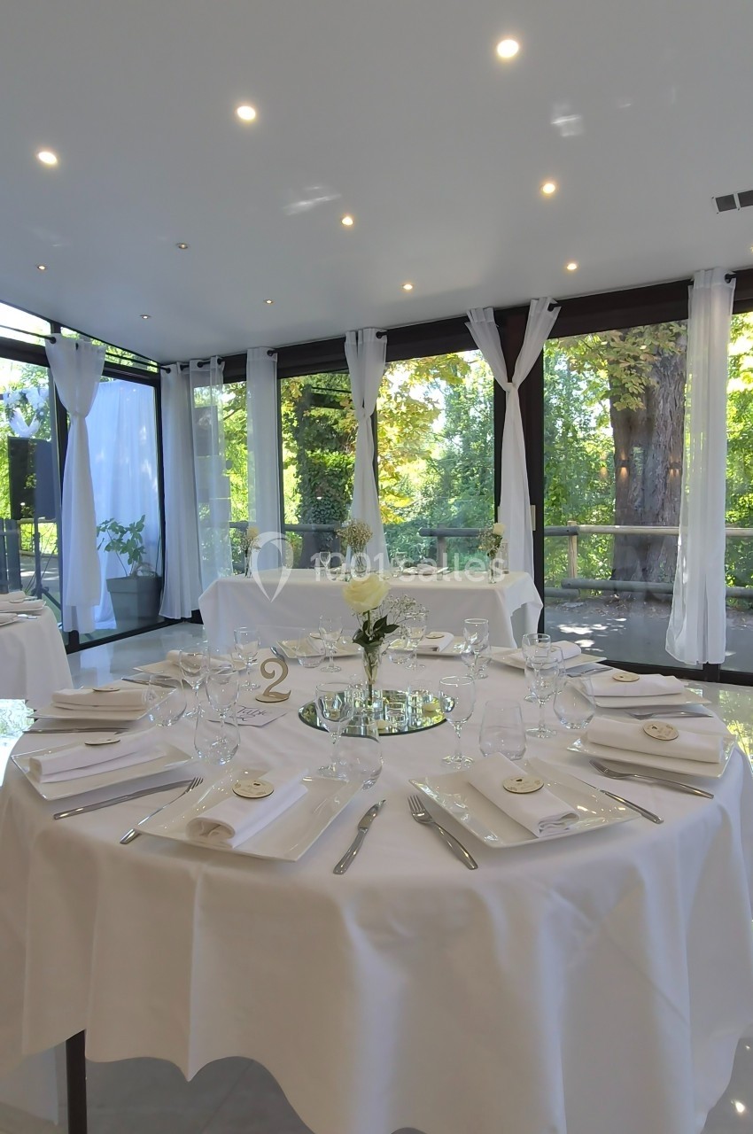 Salle de réception lumineuse avec tables rondes dressées, nappes blanches et vue sur un jardin arboré.