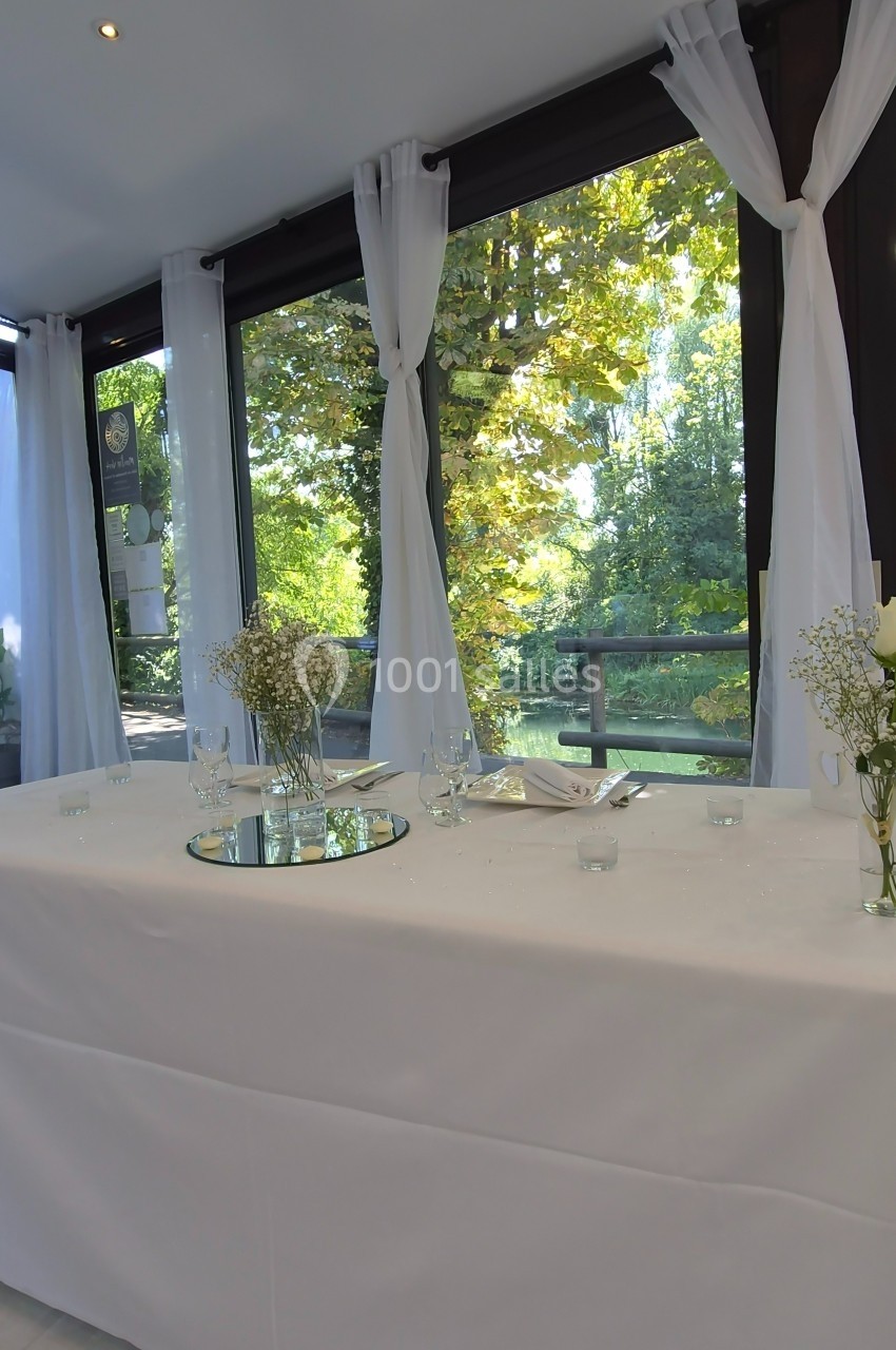 Table décorée avec nappes blanches, fleurs et bougies, placée devant de grandes baies vitrées donnant sur un paysage arboré.