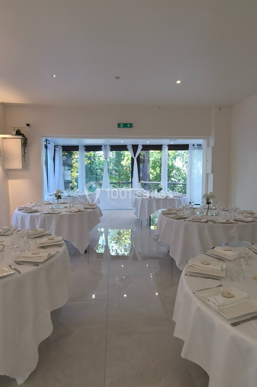 Salle de réception lumineuse avec tables rondes dressées, nappes blanches et vue sur un jardin à travers de grandes baies…