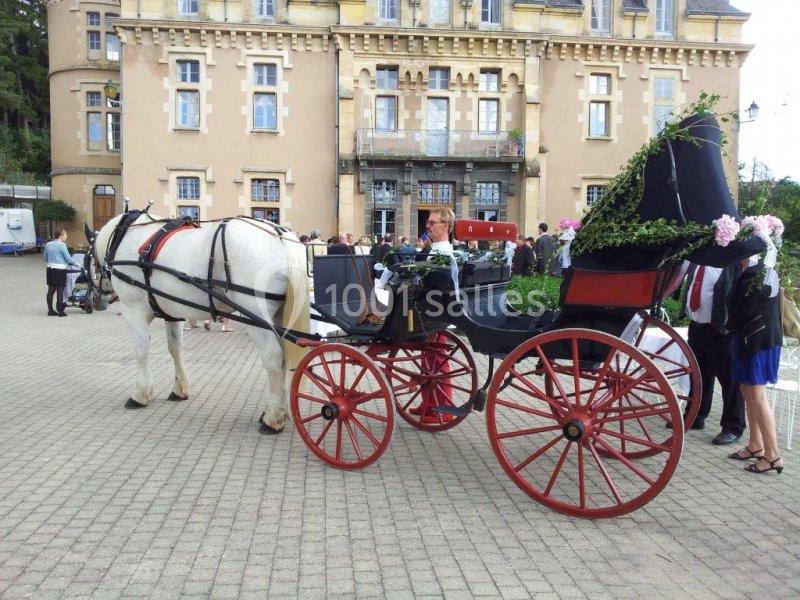 Calèche noire avec roues rouges tirée par un cheval blanc, stationnée devant un bâtiment ancien.