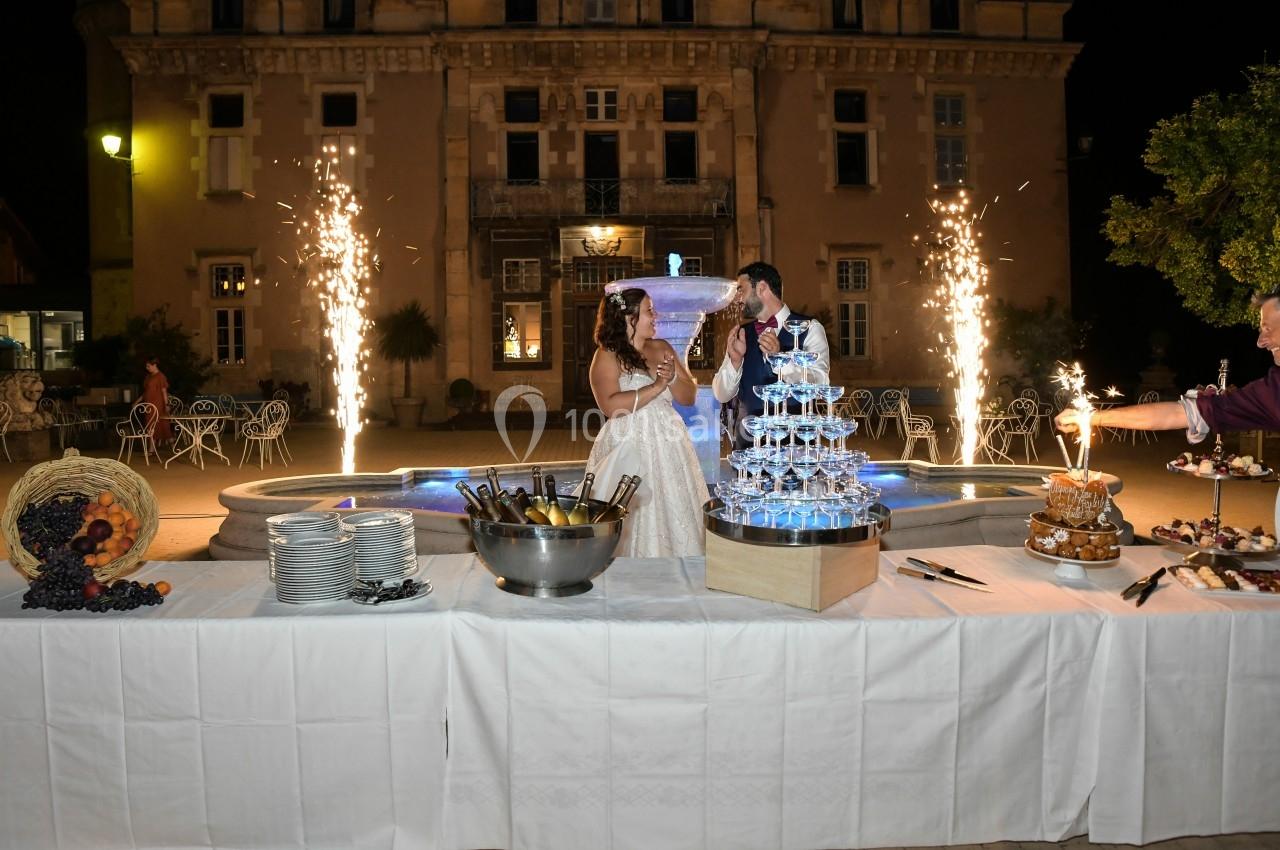Un couple célèbre son mariage devant une fontaine illuminée, avec une table garnie de desserts et de coupes de champagne.