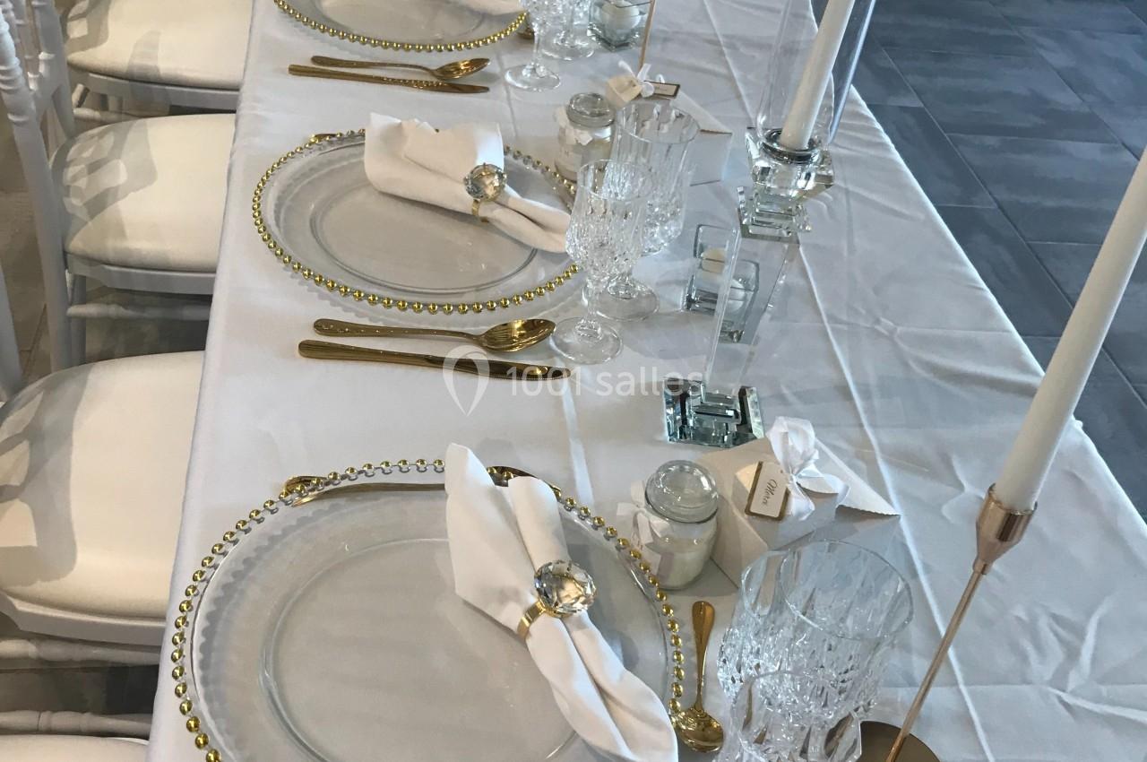 Table élégamment dressée avec assiettes transparentes, couverts dorés, serviettes pliées et chandeliers blancs.