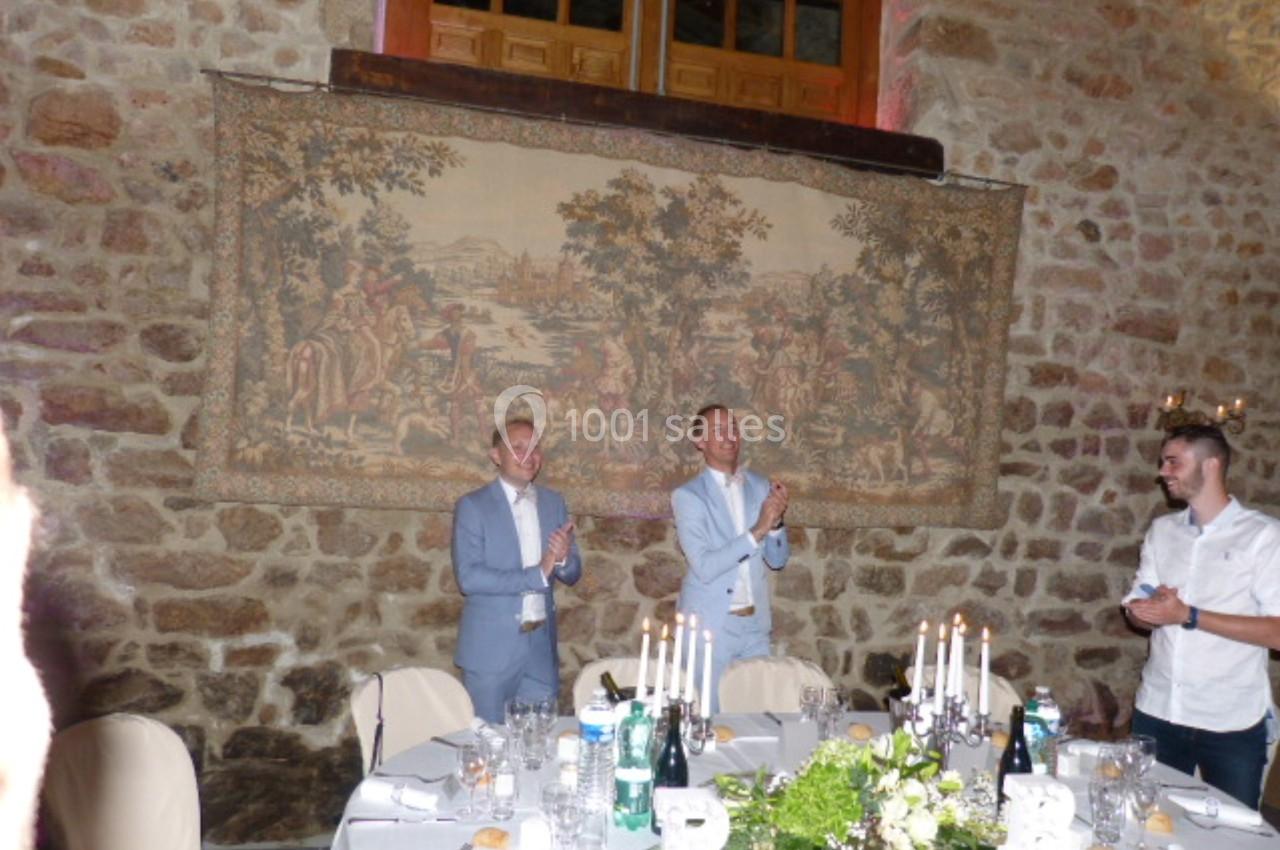 Deux hommes applaudissent debout devant une tapisserie murale dans une salle en pierre décorée pour un repas.