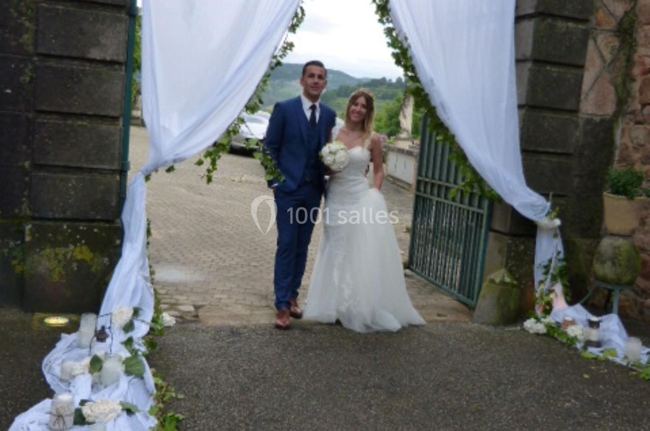 Un couple en tenue de mariage pose sous une arche décorée de drapés blancs et de fleurs, à l'extérieur.