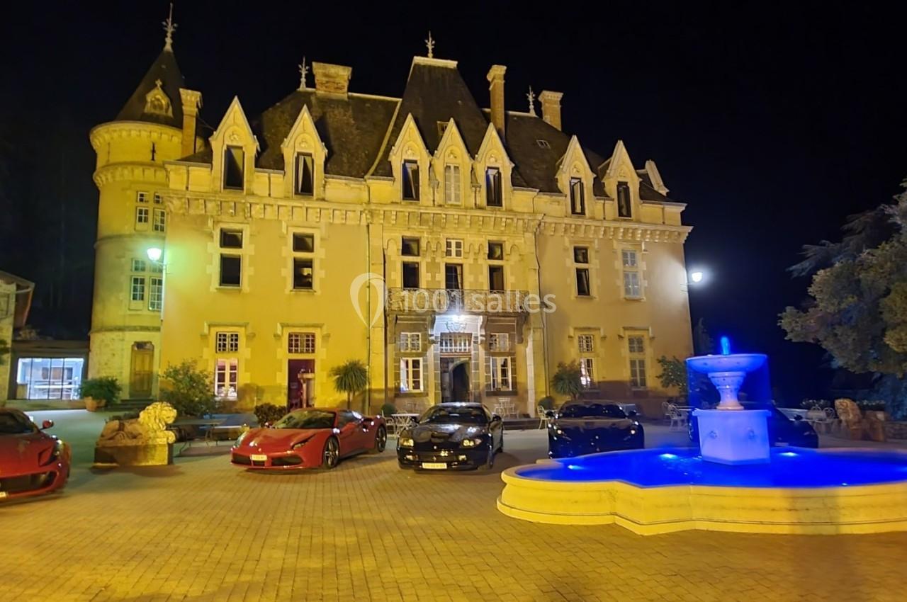 Façade éclairée d'un château de nuit avec une fontaine illuminée au premier plan et des voitures garées devant.
