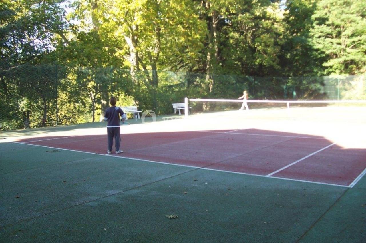 Un enfant joue sur un court de tennis extérieur entouré d'arbres, avec une autre personne en arrière-plan.