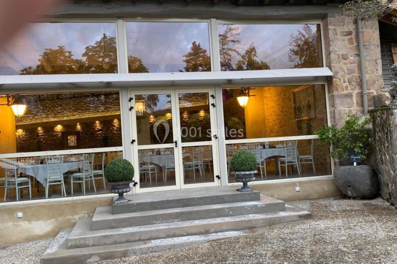 Entrée vitrée d'un bâtiment en pierre avec des tables dressées visibles à l'intérieur et des arbustes décoratifs devant.