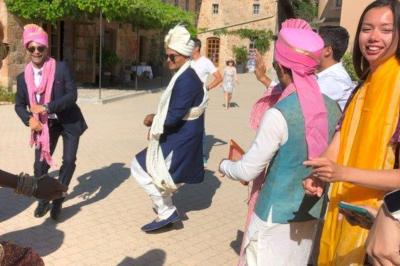 Des personnes en tenue traditionnelle dansent et célèbrent en extérieur, devant des bâtiments en pierre et végétation.