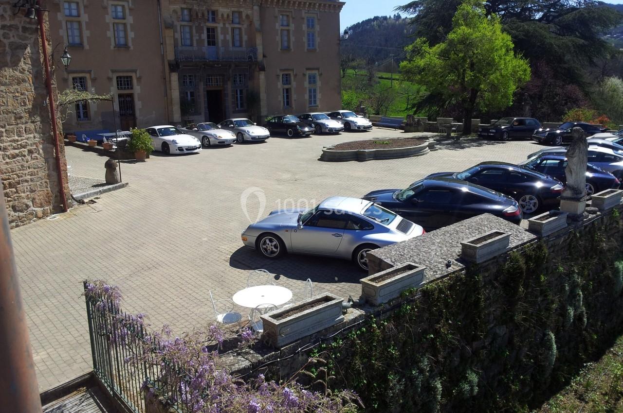 Plusieurs voitures de sport garées sur une cour pavée devant un bâtiment ancien entouré de verdure.