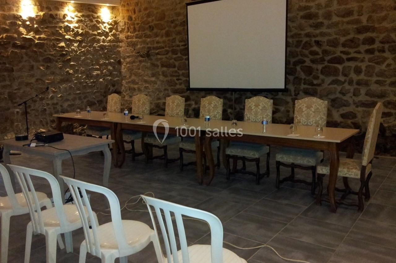 Salle en pierre avec une table longue, chaises alignées, un écran de projection et des chaises en plastique au premier plan.