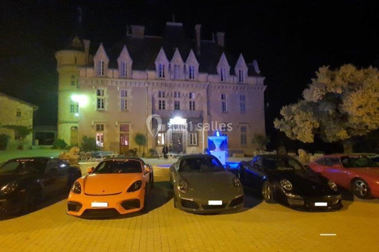 Cinq voitures de sport alignées devant un château éclairé la nuit, avec une fontaine illuminée au centre.