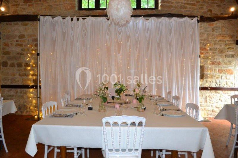 Table décorée pour un repas, avec nappes blanches, chaises assorties, fleurs et rideau lumineux en arrière-plan.