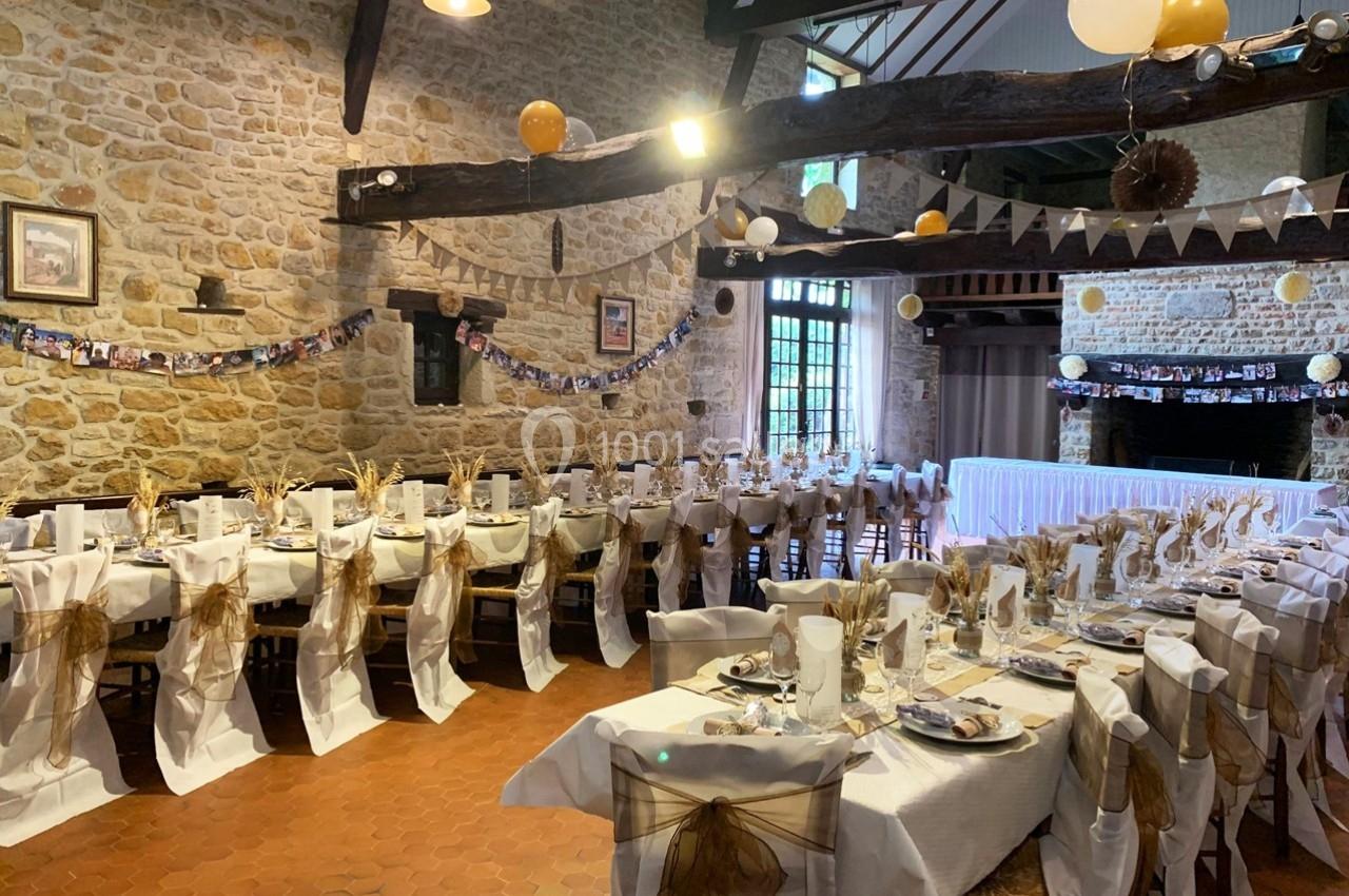 Salle de réception décorée pour un événement, avec longues tables dressées, nappes blanches et chaises ornées de nœuds dorés.