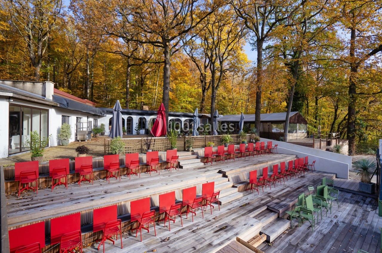 Terrasse en bois avec des rangées de chaises rouges et vertes, entourée d'arbres aux feuilles automnales.