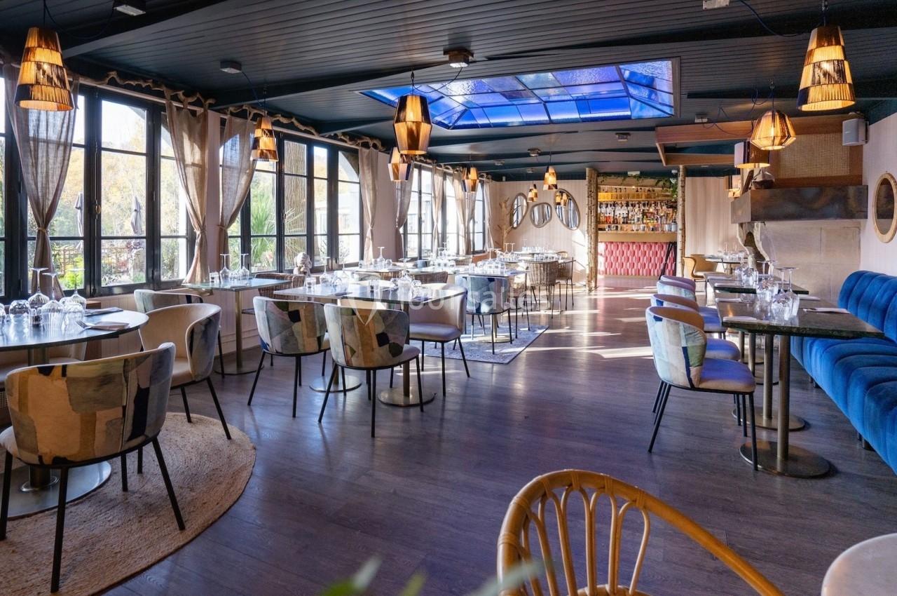 Salle de restaurant lumineuse avec grandes baies vitrées, mobilier moderne et bar en arrière-plan.