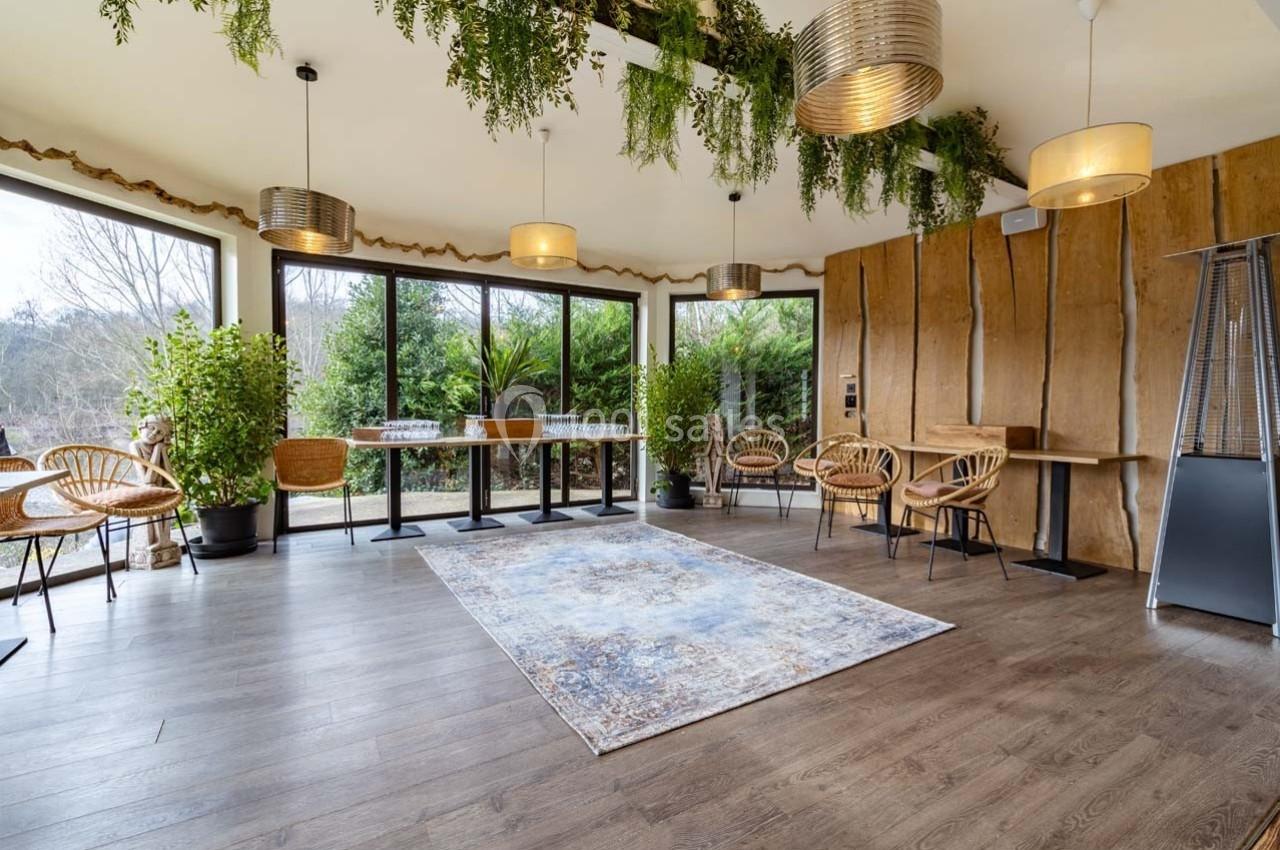 Salle lumineuse avec de grandes baies vitrées, plantes suspendues, mobilier en bois et tapis au centre.