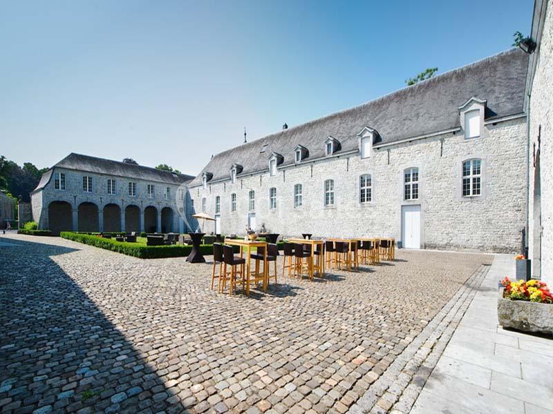 Cour pavée avec des bâtiments en pierre, tables hautes disposées en extérieur et ciel dégagé en arrière-plan.