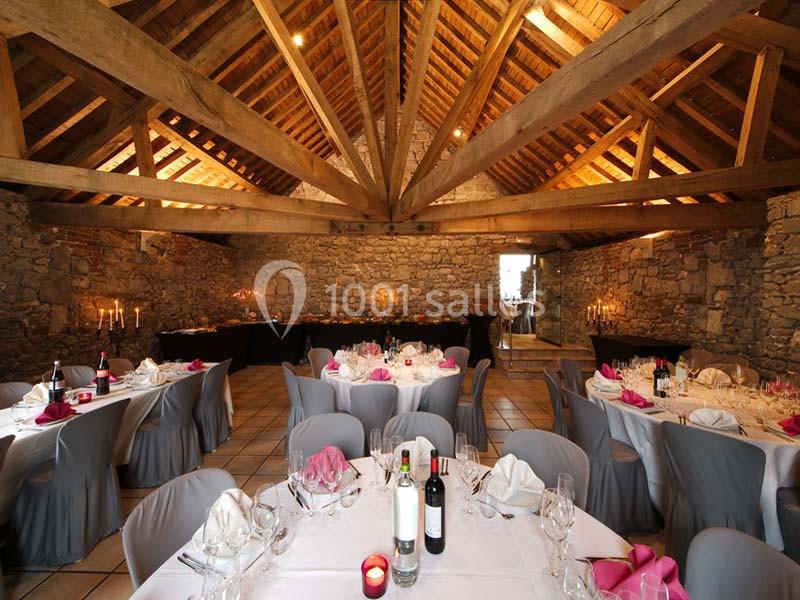 Salle en pierre avec charpente apparente, tables rondes dressées avec nappes grises et décorations roses.