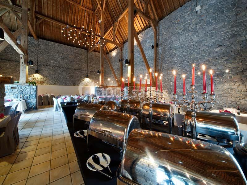 Salle de réception avec murs en pierre, poutres apparentes, chandeliers rouges et buffet en inox.
