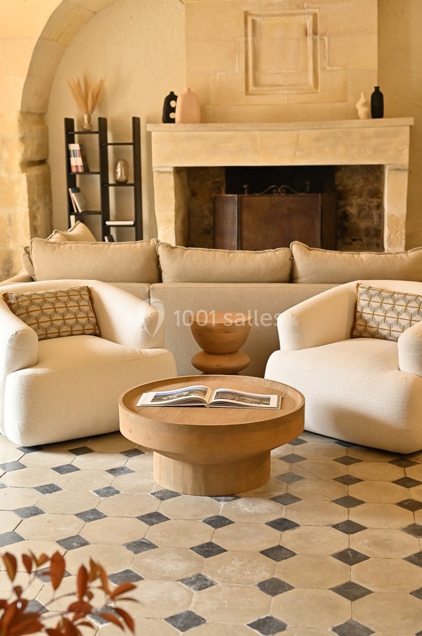 Salon chaleureux avec cheminée en pierre, canapé beige, fauteuils blancs, table basse en bois et sol en carreaux.