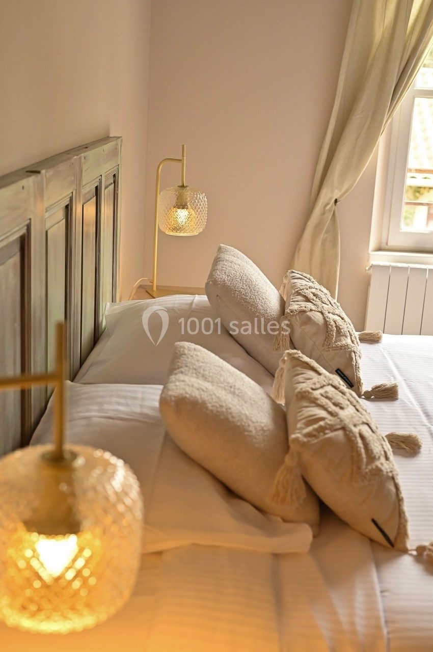 Lit double avec coussins beiges et lampes de chevet allumées, dans une chambre lumineuse avec rideaux clairs.