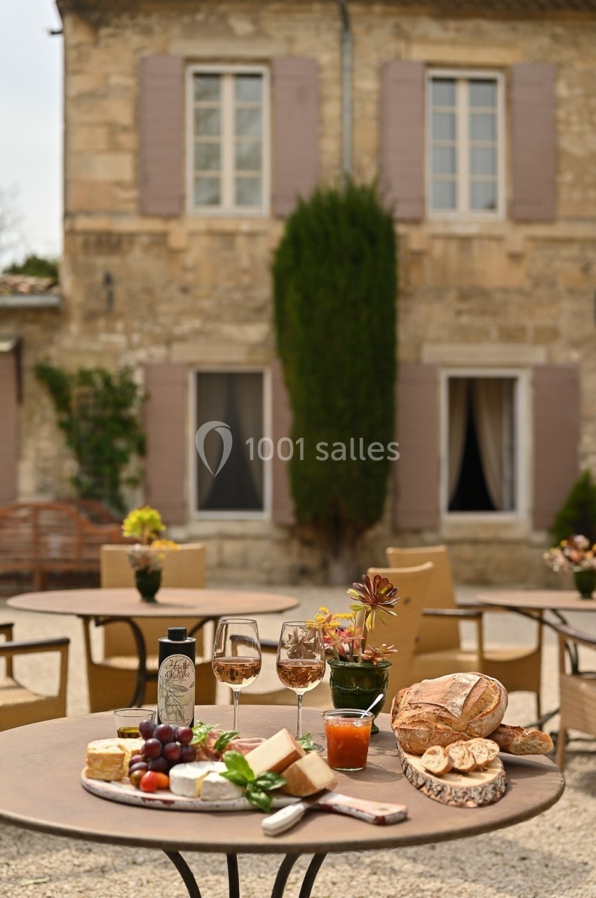 Table dressée en extérieur avec vin, pain, fromage et fruits devant une maison en pierre aux volets mauves.