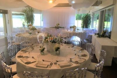 Salle de réception décorée pour un mariage, avec des tables rondes dressées, nappes blanches et arrangements floraux.