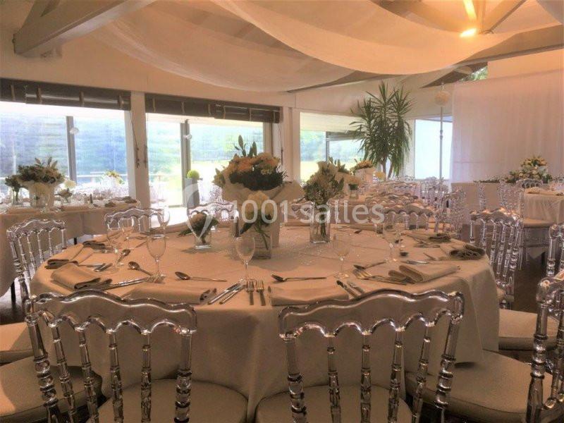 Salle de réception lumineuse avec tables rondes dressées, nappes blanches et chaises transparentes.
