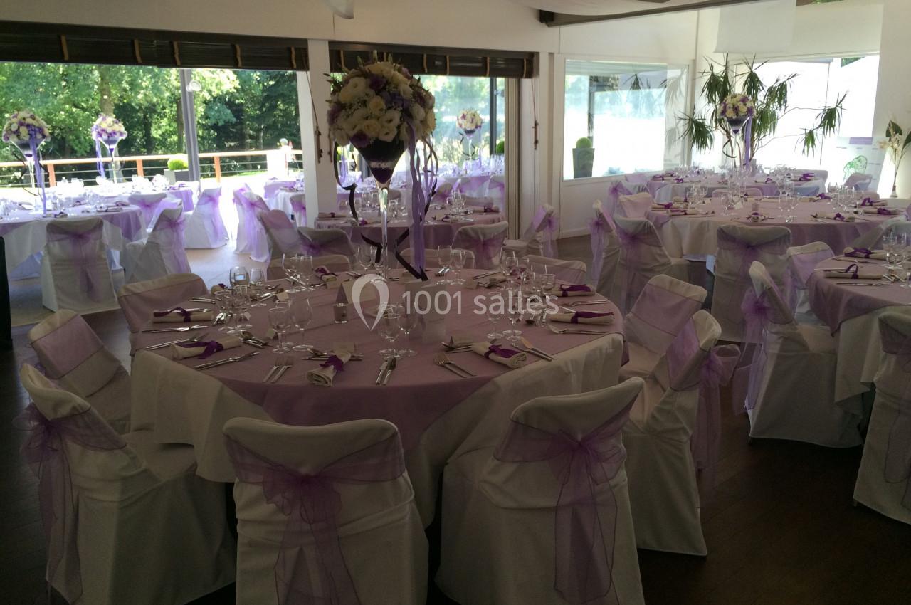 Salle de réception décorée pour un événement, avec des tables rondes, nappes blanches et ornements violets.