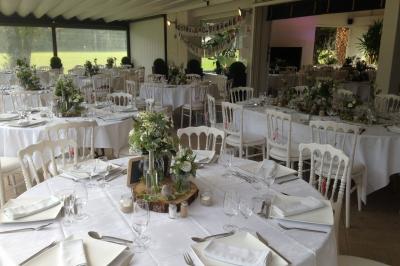 Salle de réception décorée pour un mariage, avec des tables rondes dressées, nappes blanches et arrangements floraux.