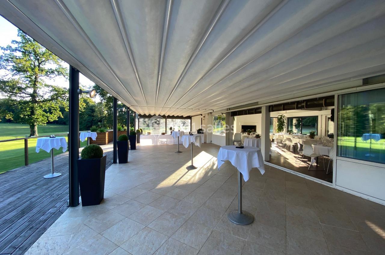 Terrasse couverte avec tables hautes dressées, vue sur un jardin verdoyant et grandes baies vitrées.