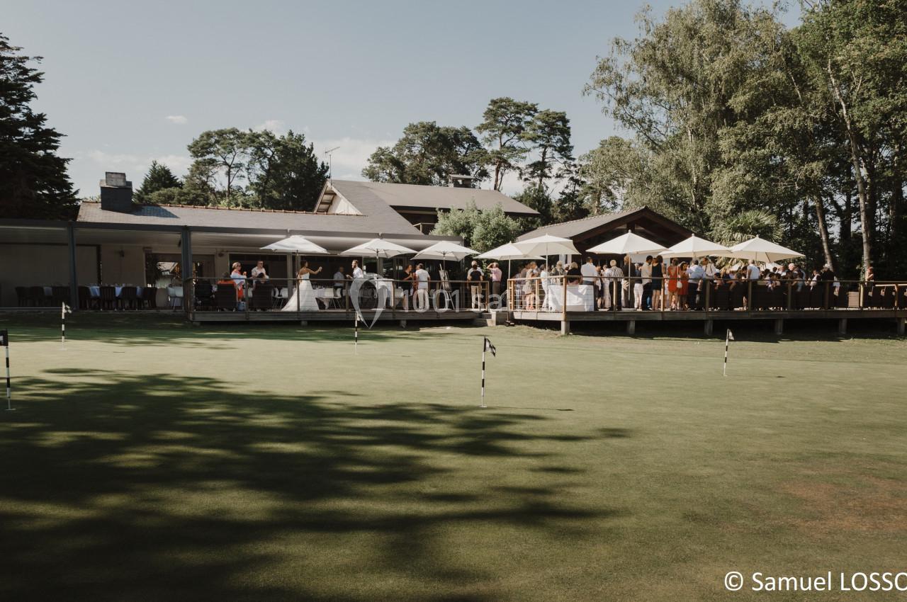 Terrasse d'un bâtiment avec des parasols et des convives, vue depuis un terrain de golf sous un ciel dégagé.