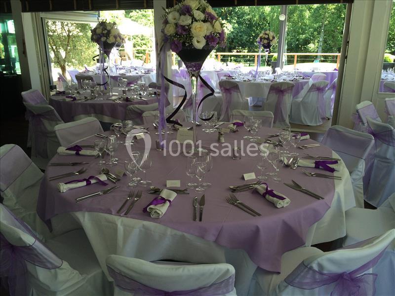 Salle de réception décorée avec des tables rondes, nappes violettes, chaises blanches et arrangements floraux.