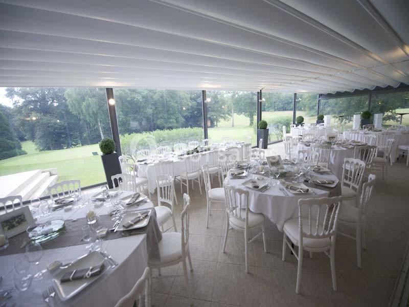 Salle de réception lumineuse avec tables rondes dressées, chaises blanches et vue sur un jardin verdoyant à travers de…