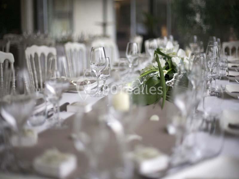 Table élégamment dressée avec nappes blanches, verres, couverts et centre de table floral dans un cadre lumineux.