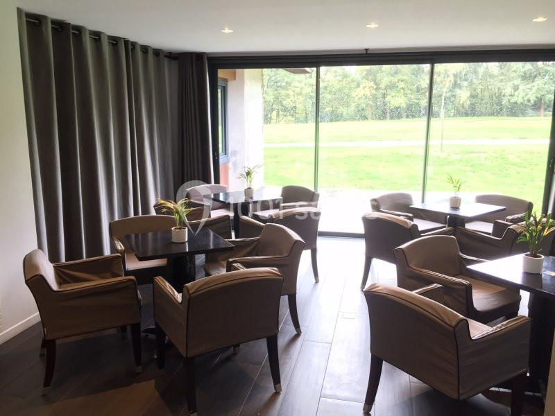 Salle lumineuse avec des tables et des fauteuils, vue sur un espace vert à travers de grandes baies vitrées.