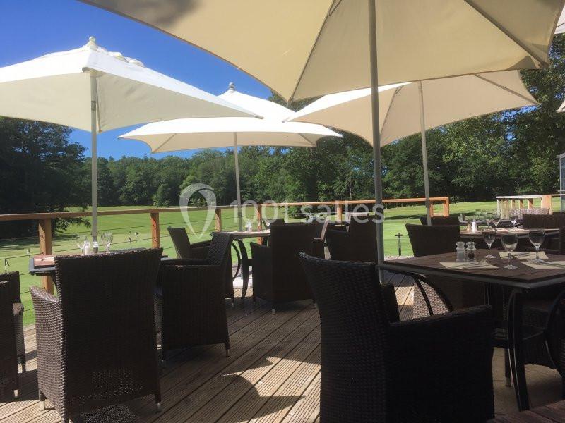 Terrasse en bois avec tables, chaises en rotin et parasols blancs, donnant sur un espace vert arboré.